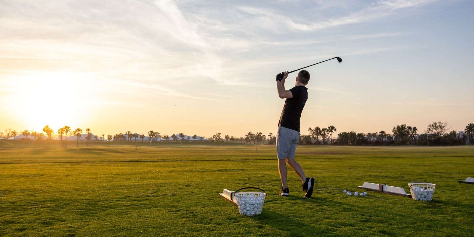 A golfer mid-swing on a grassy field at sunset with buckets of golf balls nearby.